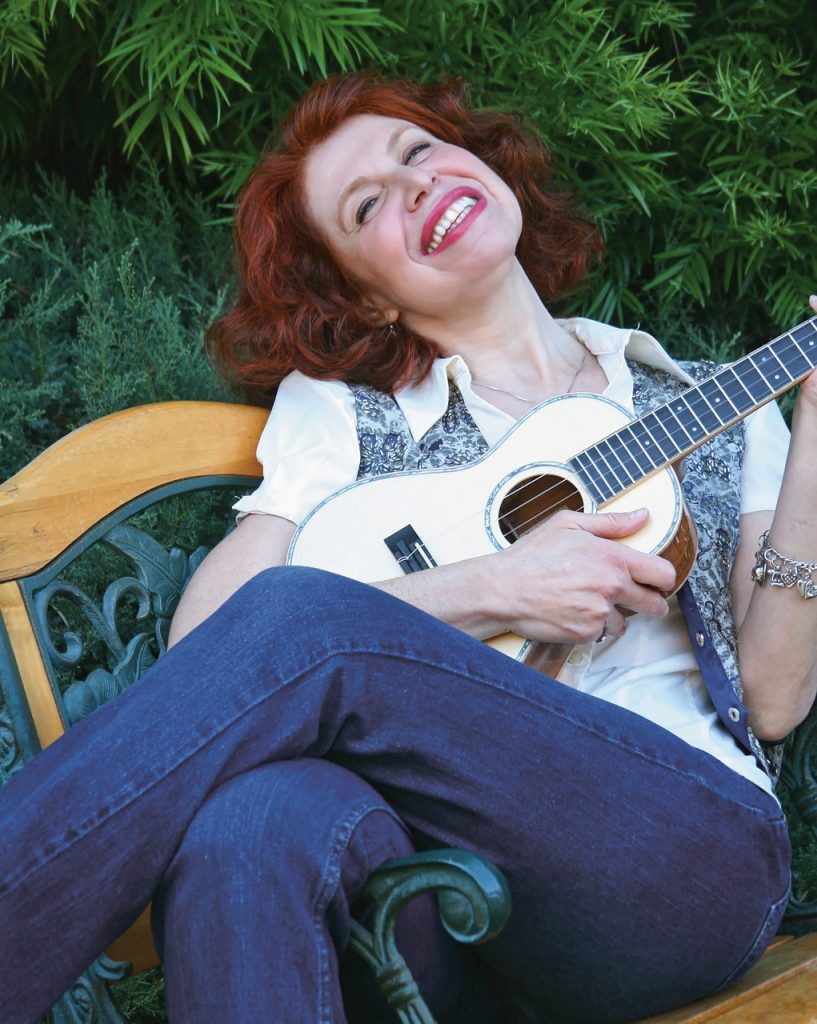 A red-haired woman with a ukulele reclines and smiles on a bench outdoors.