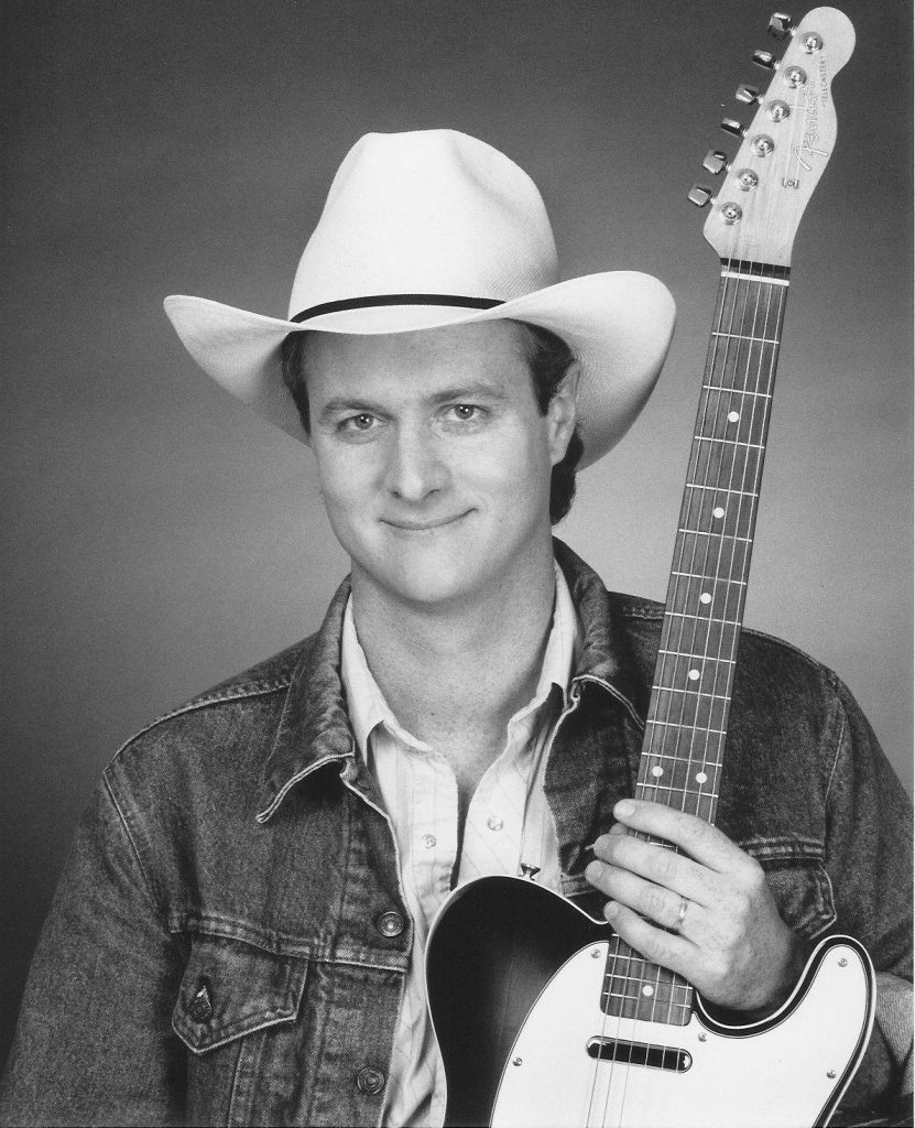 A smiling young man in a cowboy hat holding a guitar