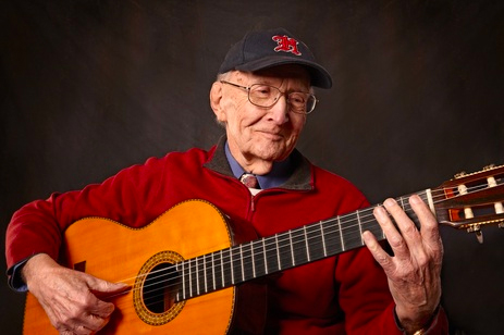 An older man in a baseball hat and red sweater plays an acoustic guitar.